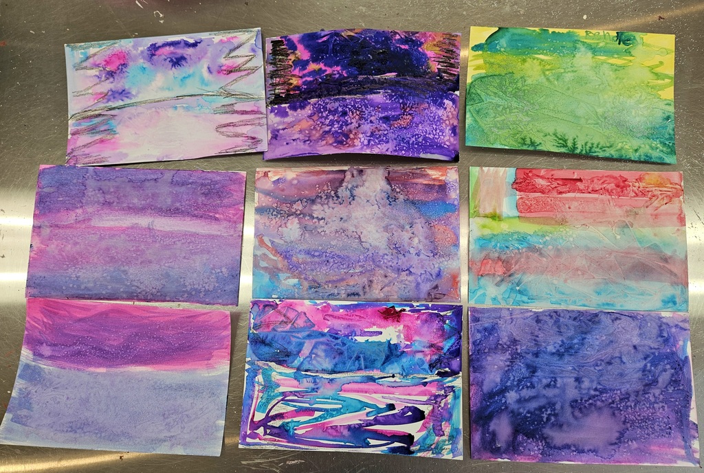 A collection of nine student watercolor paintings laid out on a table. The paintings feature vibrant blues, purples, and pinks with visible textures from salt and saran wrap to create 'burst' and 'ripple' effects.