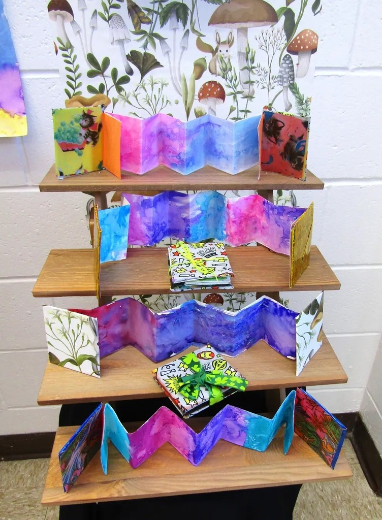 A three-tiered wooden display stand showcasing several handmade student books. The books feature colorful accordion-folded watercolor pages inside and a variety of decorative covers including floral patterns, kittens, and bright illustrations.