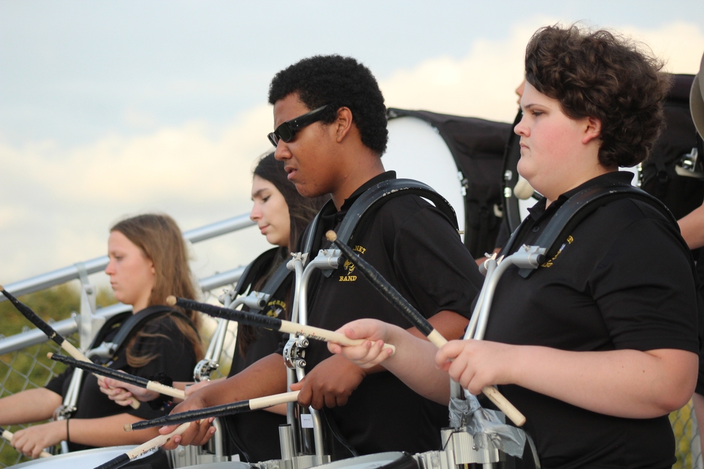 Students in the percussion group of the band play at a football game. All wearing black polos and drumming to the beat and focused.