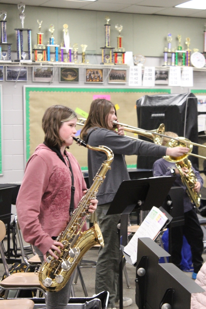 Two students play their instruments in band class, a saxophone and a trombone. Trophies line the walls behind them. 