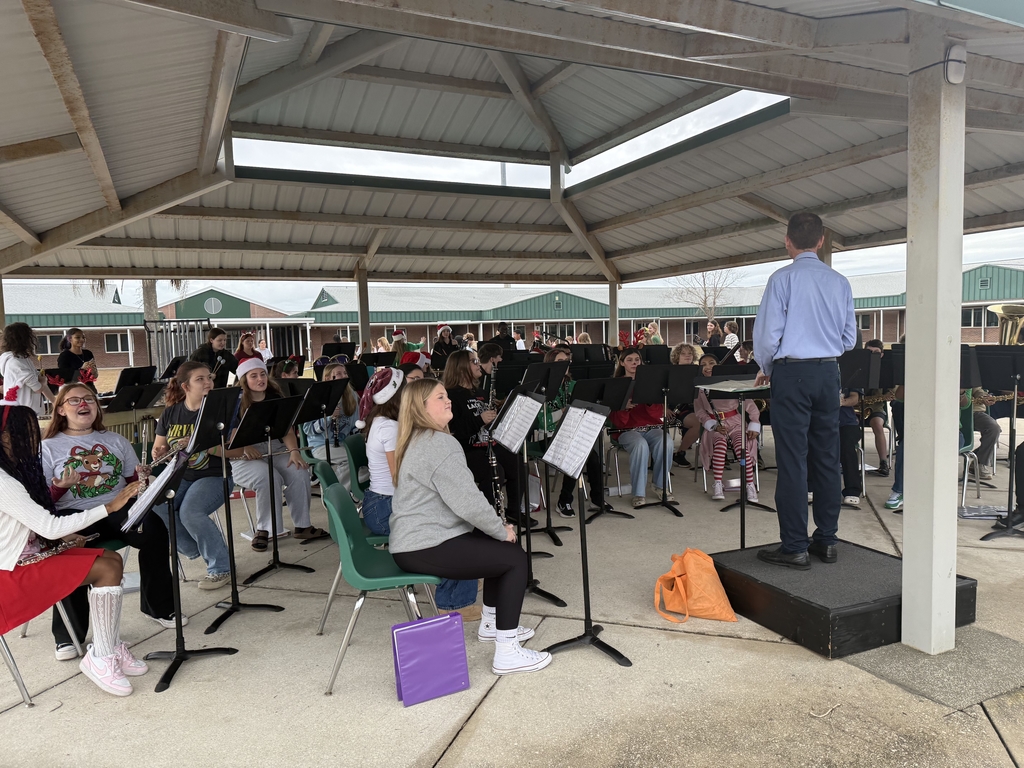 Band Director stands at the front of students who are seated and watch for him to direct their next song. They are under a pavilion.