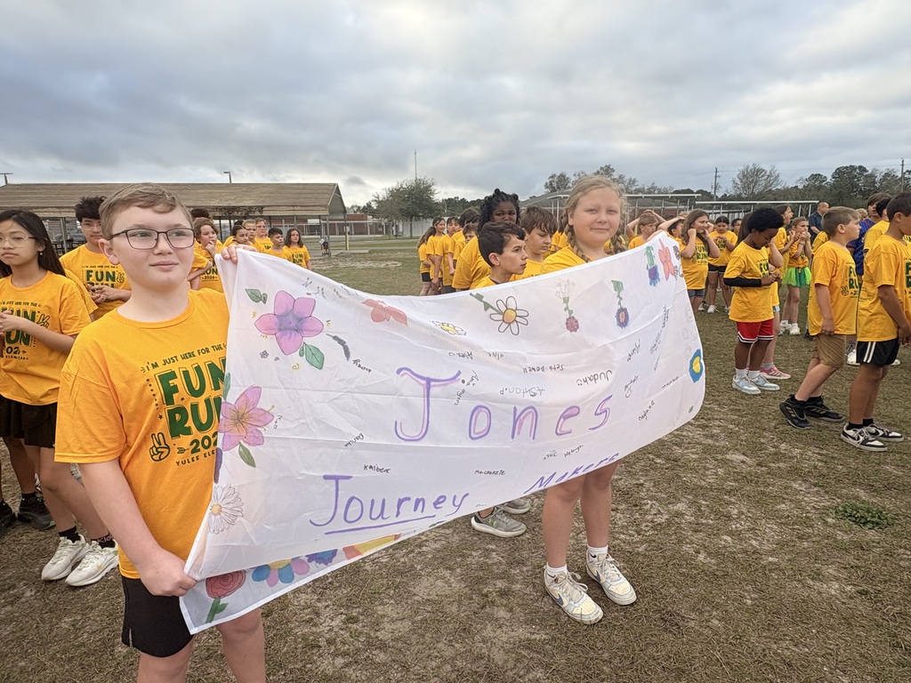 Two elementary students in yellow Hornet t-shirts holding up a large, hand-decorated white banner that says 'Jones Journey' with colorful musical notes and drawings. Other students are lined up in the background on the school field.
