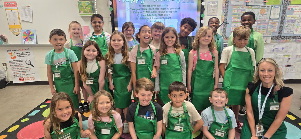 A large group of 2nd-grade students and their teacher at Wildlight Elementary, all wearing green barista-style aprons and smiling in front of a classroom whiteboard during their Starbucks-themed Poetry Week.