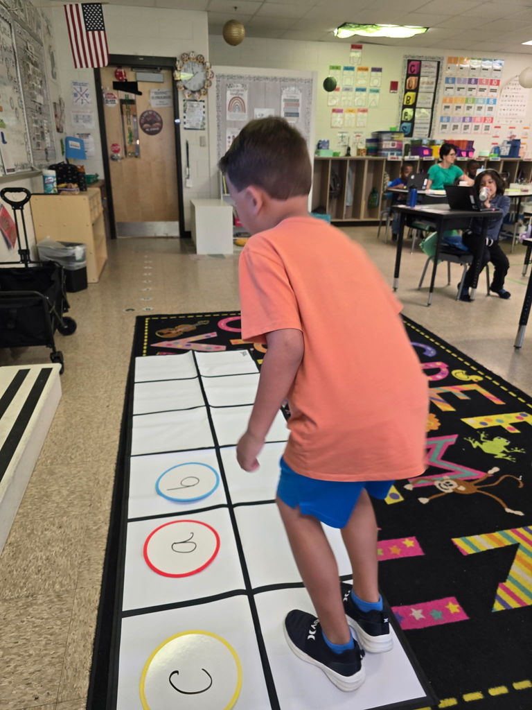 Student at Wildlight Elementary are participating in an interactive phonics lesson. They are jumping along a large floor mat printed with letters and words, physically sounding out each step as they move through the activity in a bright, modern classroom.