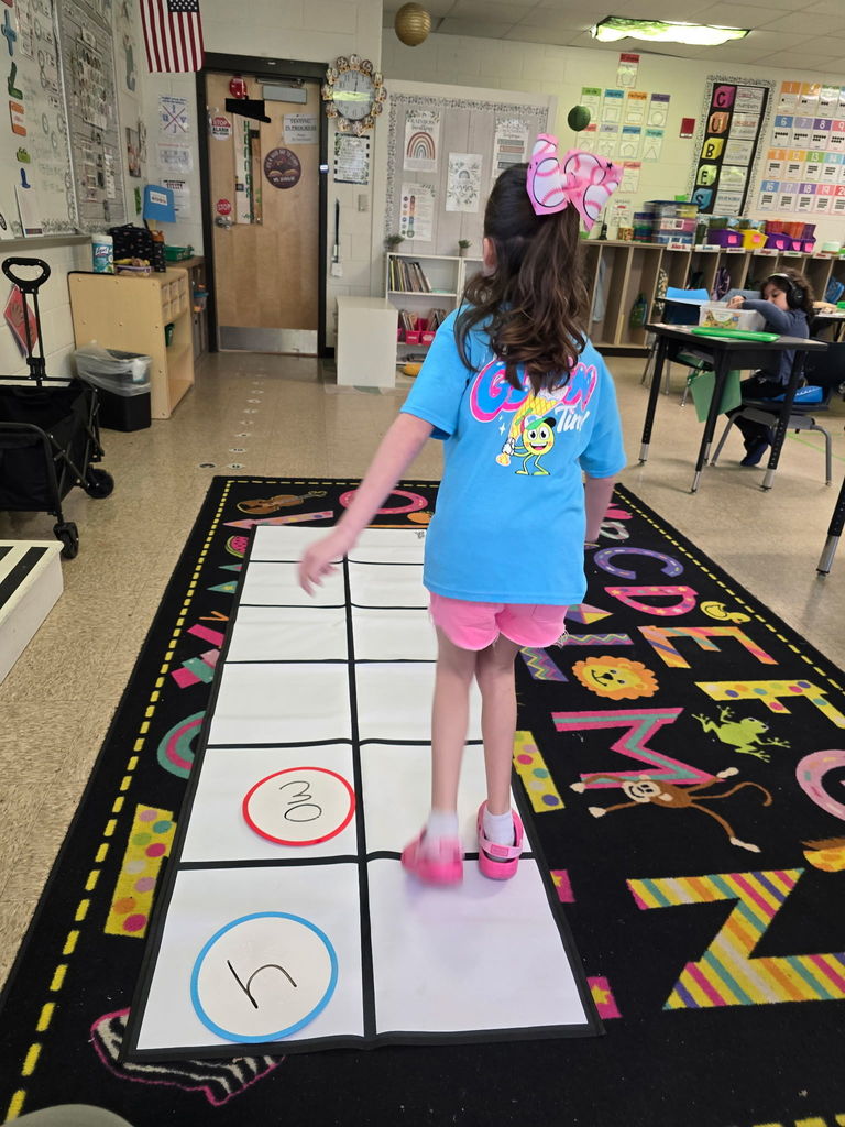 Student at Wildlight Elementary are participating in an interactive phonics lesson. They are jumping along a large floor mat printed with letters and words, physically sounding out each step as they move through the activity in a bright, modern classroom.