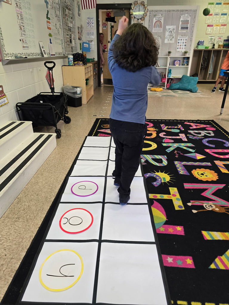 Student at Wildlight Elementary are participating in an interactive phonics lesson. They are jumping along a large floor mat printed with letters and words, physically sounding out each step as they move through the activity in a bright, modern classroom.