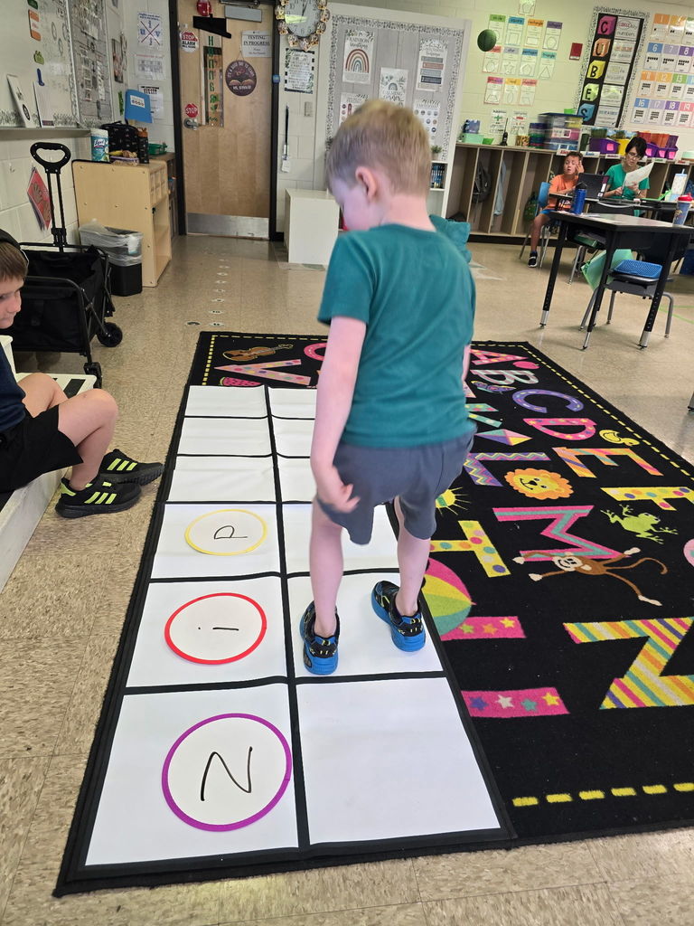 Student at Wildlight Elementary are participating in an interactive phonics lesson. They are jumping along a large floor mat printed with letters and words, physically sounding out each step as they move through the activity in a bright, modern classroom.