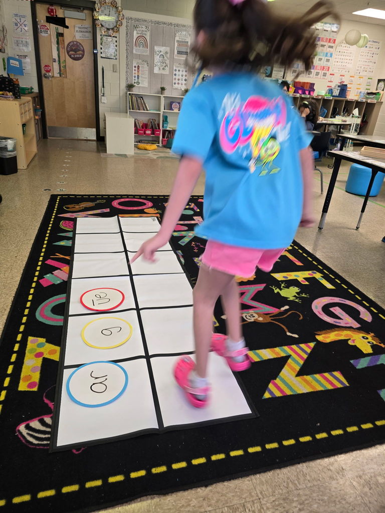 Student at Wildlight Elementary are participating in an interactive phonics lesson. They are jumping along a large floor mat printed with letters and words, physically sounding out each step as they move through the activity in a bright, modern classroom.