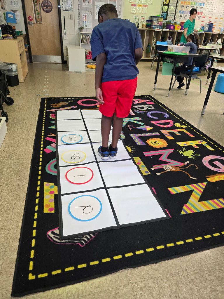 Student at Wildlight Elementary are participating in an interactive phonics lesson. They are jumping along a large floor mat printed with letters and words, physically sounding out each step as they move through the activity in a bright, modern classroom.
