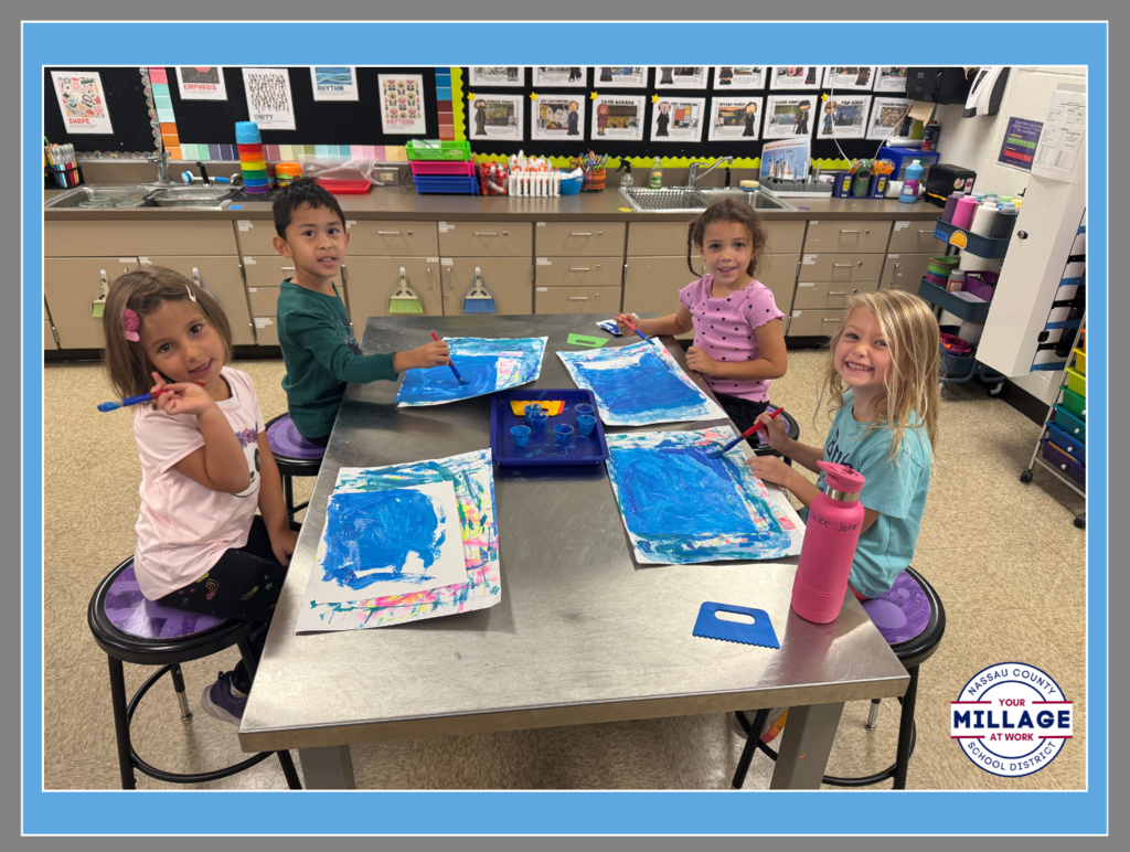 Four elementary students smiling while working on a blue painting project at a classroom table.