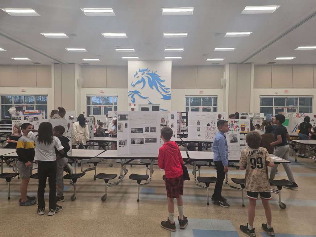 A wide shot of the Wildlight Elementary cafeteria transformed into a Living Museum, with 4th-grade students standing at rows of tables presenting their research projects to visitors.