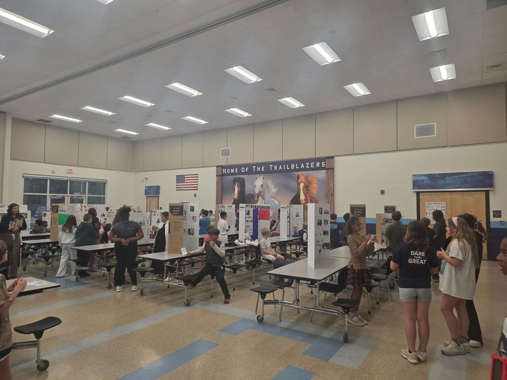 A wide shot of the Wildlight Elementary cafeteria transformed into a Living Museum, with 4th-grade students standing at rows of tables presenting their research projects to visitors.