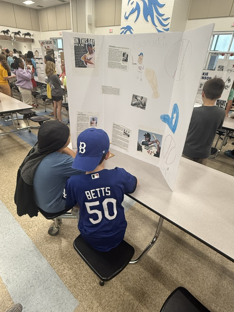 A 4th-grade student wearing a "Betts 50" jersey sits at a desk, presenting a Living Museum project to a peer. The display board is filled with research and photos about a historical figure.