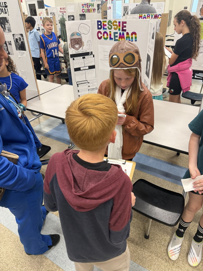 A 4th-grade student dressed as aviator Bessie Coleman, wearing flight goggles and a brown jacket, explains her research to a younger student during the Living Museum.