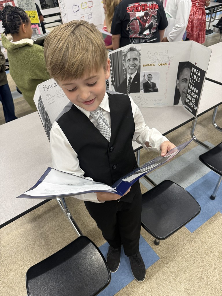 A 4th-grade student dressed in a black suit and vest smiles while holding his research notes in front of a Living Museum display about Barack Obama.