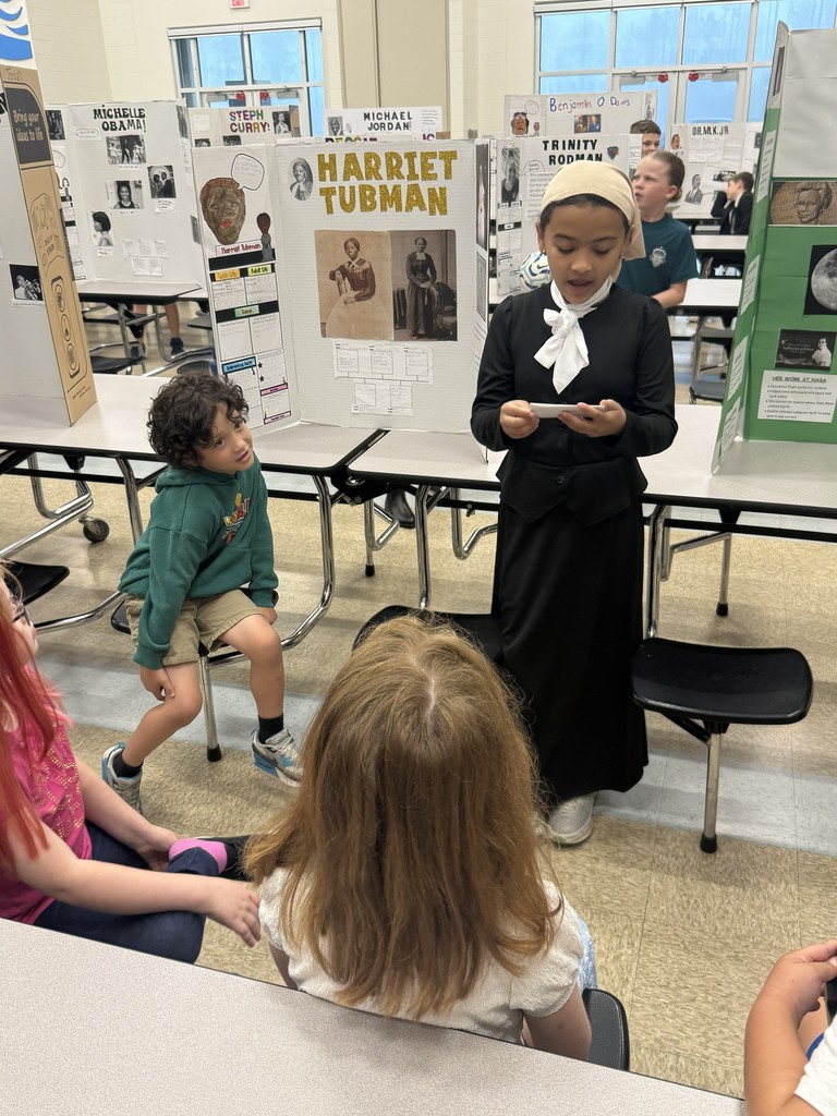 A 4th-grade student standing proudly in front of her colorful "Living Museum" display board, which features research, photos, and a timeline of a historical figure.
