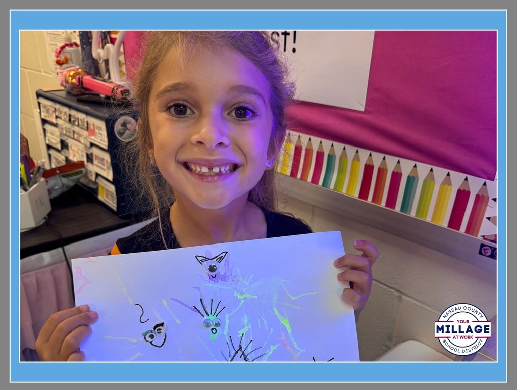 Close-up of a young girl with a big smile holding up her colorful drawing of animals in a classroom. A "Millage at Work" logo is visible in the bottom right corner.