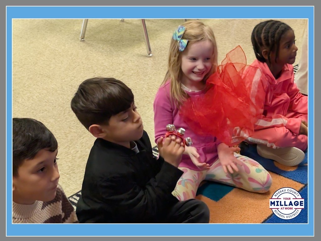 Three young students sitting together on a classroom floor, smiling and holding a red mesh fabric. A "Millage at Work" logo is visible in the bottom right corner of the frame.