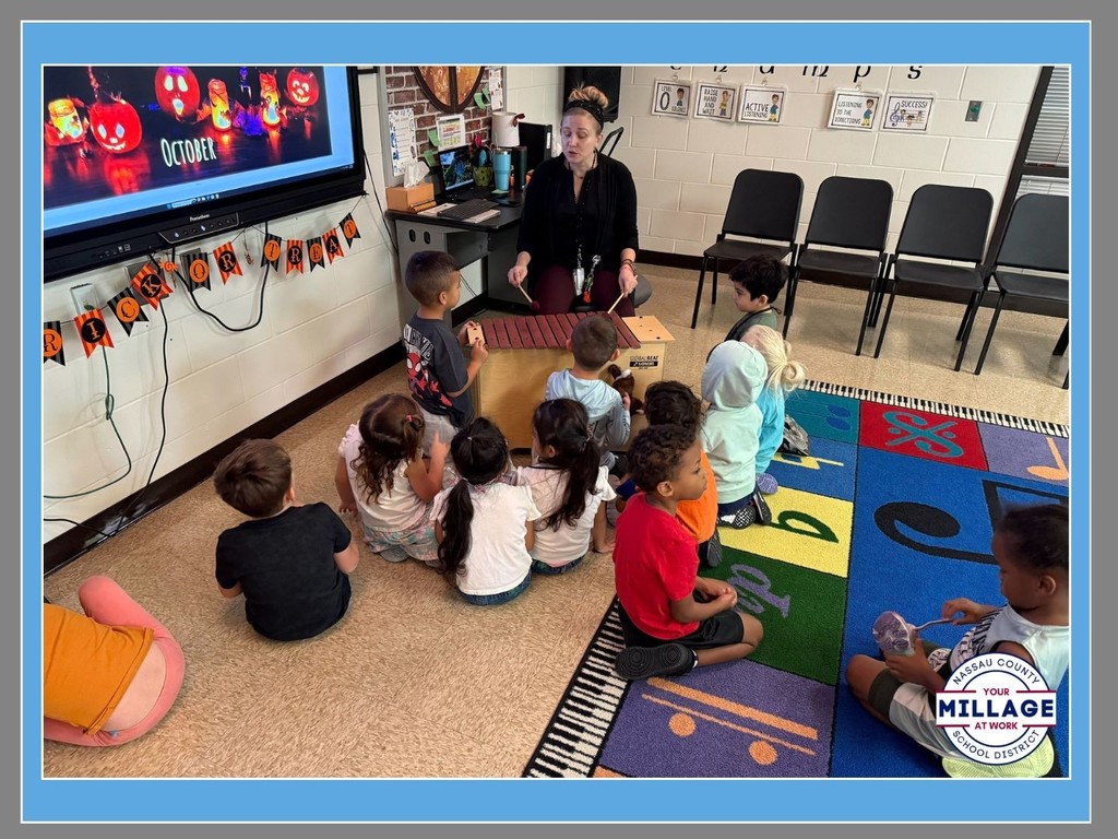 A teacher sitting on a colorful classroom rug leading a lesson for a group of young students. The students are sitting on the floor facing a large monitor displaying an "October" calendar. A "Millage at Work" logo is in the bottom right corner.