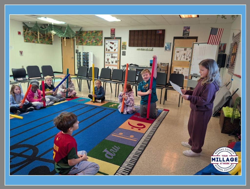 A group of elementary students sitting on a large, colorful rug in a classroom. One student stands at the front holding a paper, while others watch from the perimeter. The room is decorated with educational posters and a "Millage at Work" logo is in the bottom right corner.