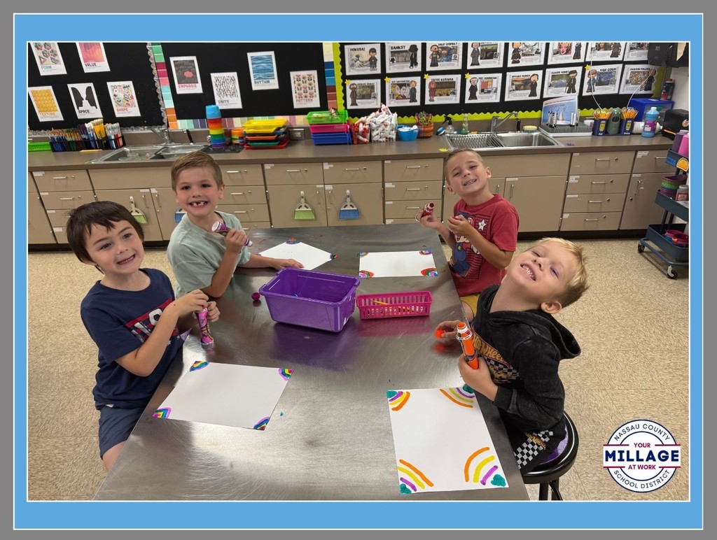 A group of elementary students smiling and working on art projects at a table in a bright classroom.