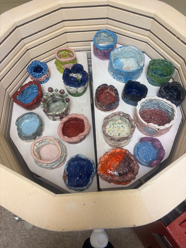 A picture of clay pottery fresh out of the kiln. There are sixteen different bowls, all made with different colors. Picture is taken from above. 