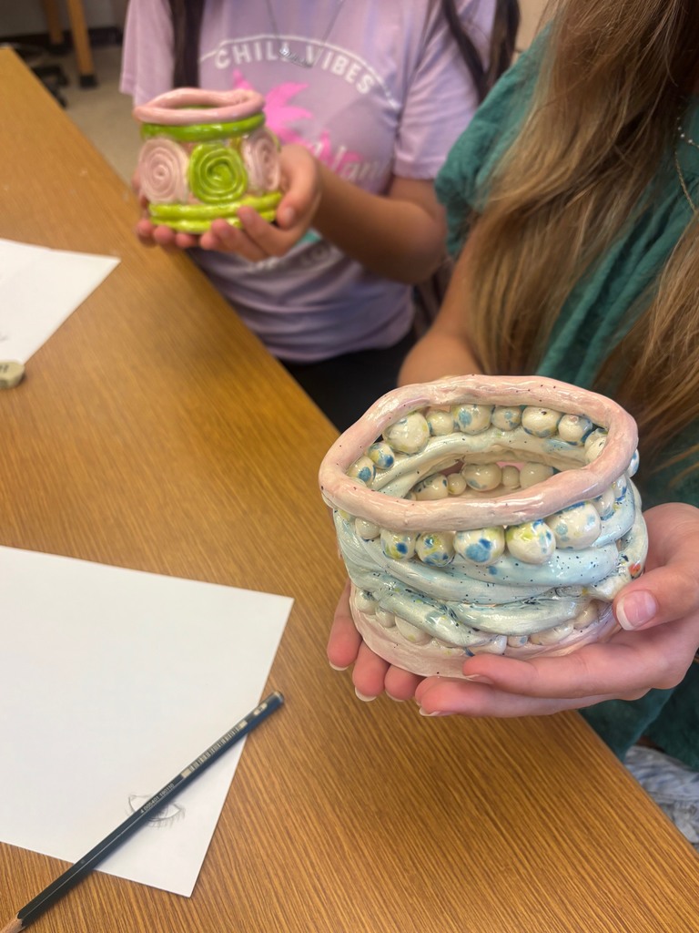 Two students show a close up of their pottery. The one closes to the camera looks like a nest with spotten yellow and blue eggs mixed in the sides of the bowl. 