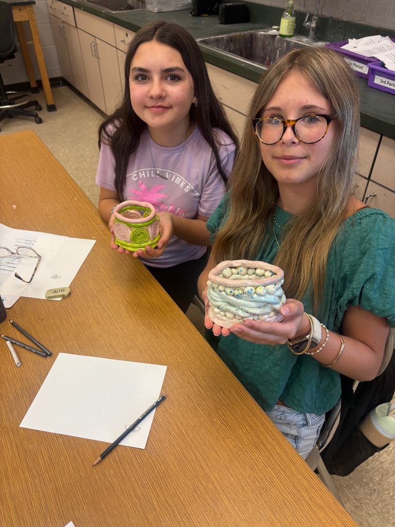 Two students hold their clay art bowls in their hands as the smile for the camera at their desk in art class.