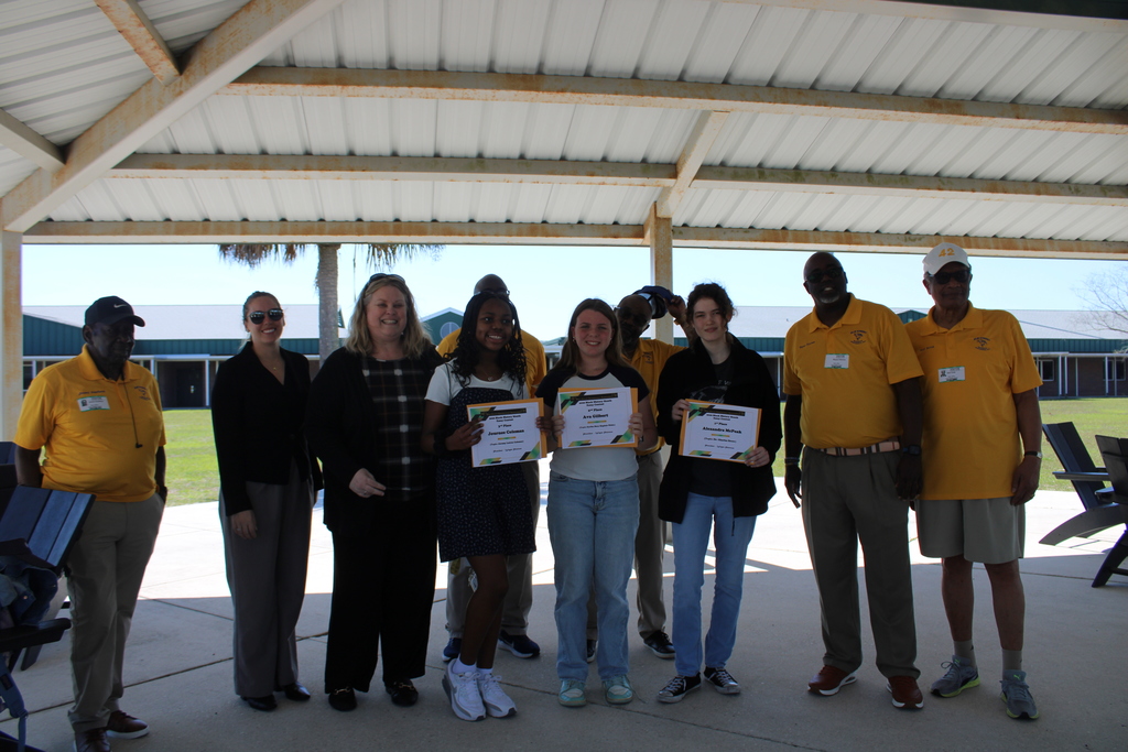 Elm Street Sportsman Association (ESSA) representatives pose with Dr. Middleton, school principal, Mrs. Miles, Journalism teacher, and the three student winners of the black history month essay contest under the school's pavilion while students hold their winning certificates.