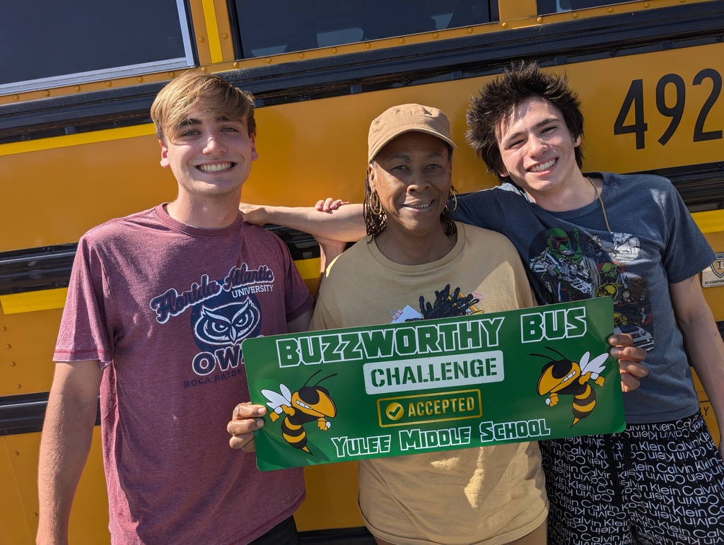 School bus driver poses with two of her student riders in front of her yellow school bus number 492. She is holding a sign that says Buzzworthy Bus, Challenge Accepted, Yulee Middle School with two hornets on either side. 