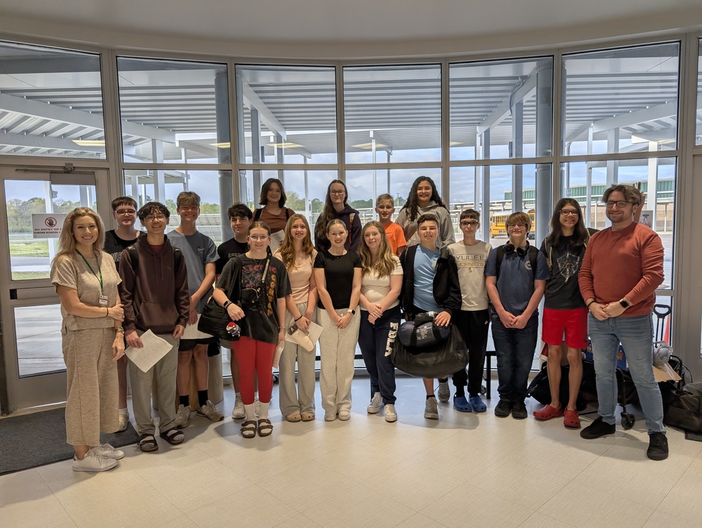 Students and their club sponsors pose and smile for the camera in front of the windows leading out to the bus parking lot  before leaving for FBLA State Finals. 