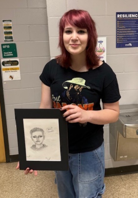 Female student with red hair smiles for the camera while holding her pencil drawing of a female student with short hair from the neck up. 