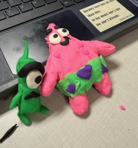 Two clay models, one of Patrick (pink with green and purple shorts) and the other of Plankton (green with one giant eye) from the show Spongebob Squarepants, created by a student lay at the base of a school laptop.