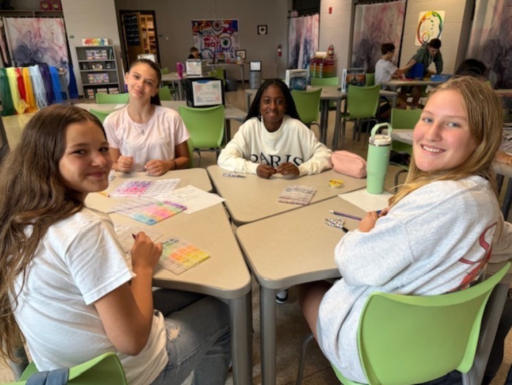 Four female students smile for the camera as they are painting at their desks in art class getting ready to do some water colors.
