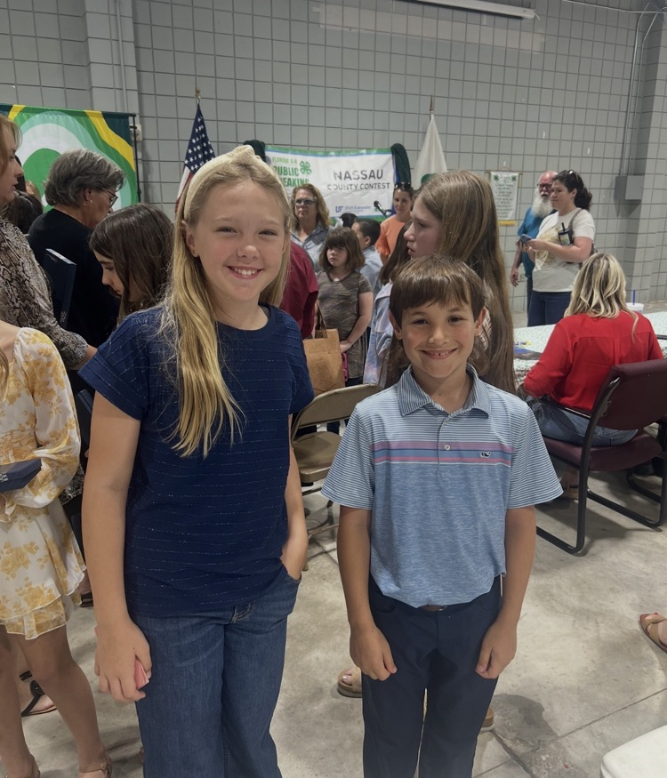 A blond g girl and a brown haired boy are standing side-by-side smiling at the camera. you can see the 4-H logo in the background. 