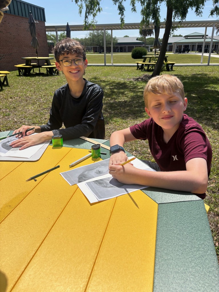 Two students smile for the camera as they work on their art projects at the new green and gold picnic tables.