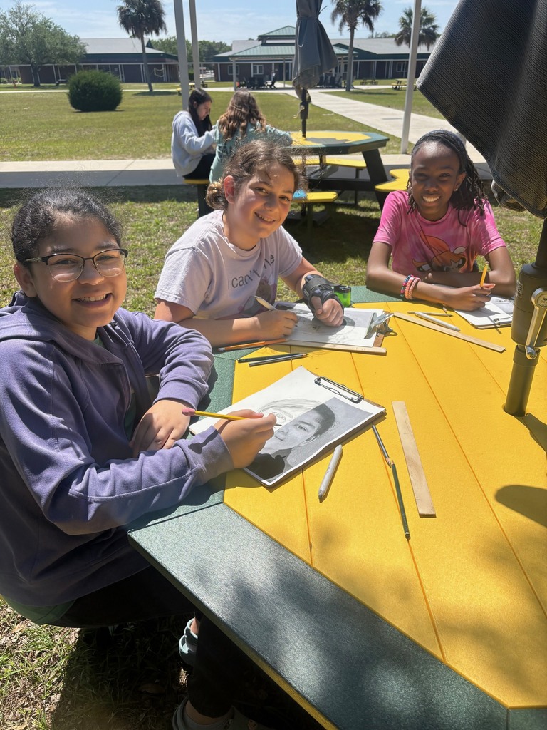 Three students smile for the camera as they work on their art projects at the new green and gold picnic tables.