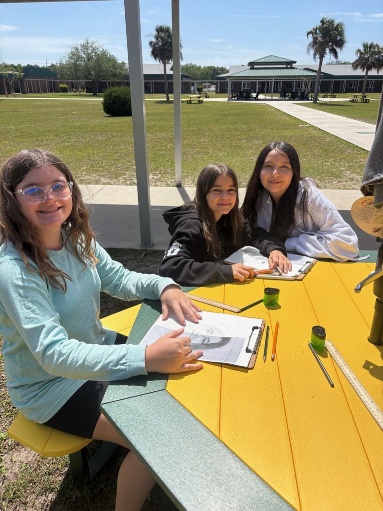 Three students, who are working on their self portraits, smile for the camera outside as they sit at the new green and gold picnic tables.