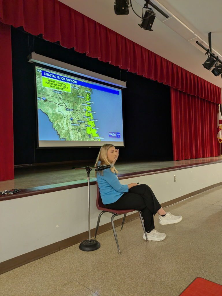 A meteorologist sits on a chair in front of a stage while presenting a weather map showing a coastal flood advisory, explaining severe weather conditions to students during a school visit.