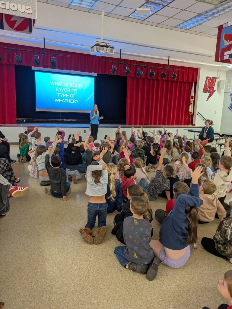 Kindergarten students sit on the floor with hands raised as a meteorologist stands on stage presenting a slide about favorite types of weather during an interactive school assembly.