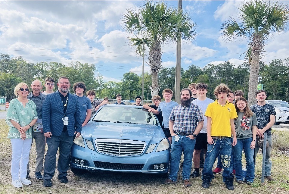 A group of Yulee High School students, staff, and community members stand outdoors around a donated Mercedes-Benz vehicle, highlighting support for the school’s automotive program and hands-on learning opportunities.