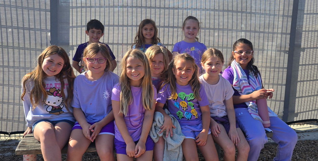 A group of nine elementary students sitting and standing together against a wire fence. They are all wearing purple shirts and smiling for a group photo to celebrate Purple Up Day.