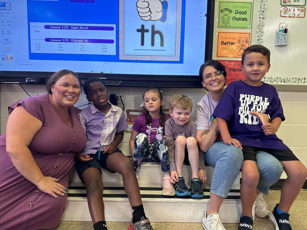 Two staff members and four elementary students sitting together in a classroom. They are all wearing purple to celebrate Purple Up Day, and one student is wearing a shirt that says "Purple Up for Military Kids."