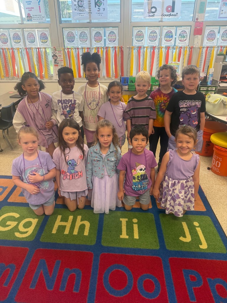 A large group of primary school students standing together on a colorful alphabet rug inside a classroom. All of the children are wearing purple shirts or dresses to celebrate Purple Up Day for military kids.