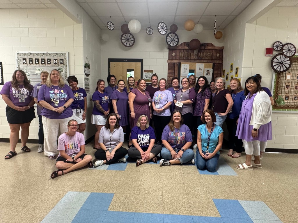 A large group of about twenty teachers and staff members posing together in a school hallway. They are all wearing various shades of purple clothing to participate in Purple Up Day for military children.
