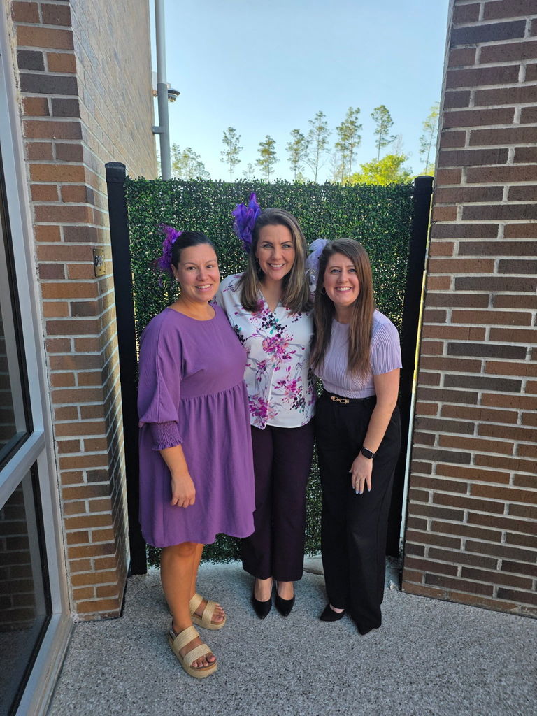 Three women standing together outdoors between two brick walls in front of a green hedge. They are all dressed in purple outfits, including a purple dress and floral blouse, to celebrate Purple Up Day at Wildlight Elementary.