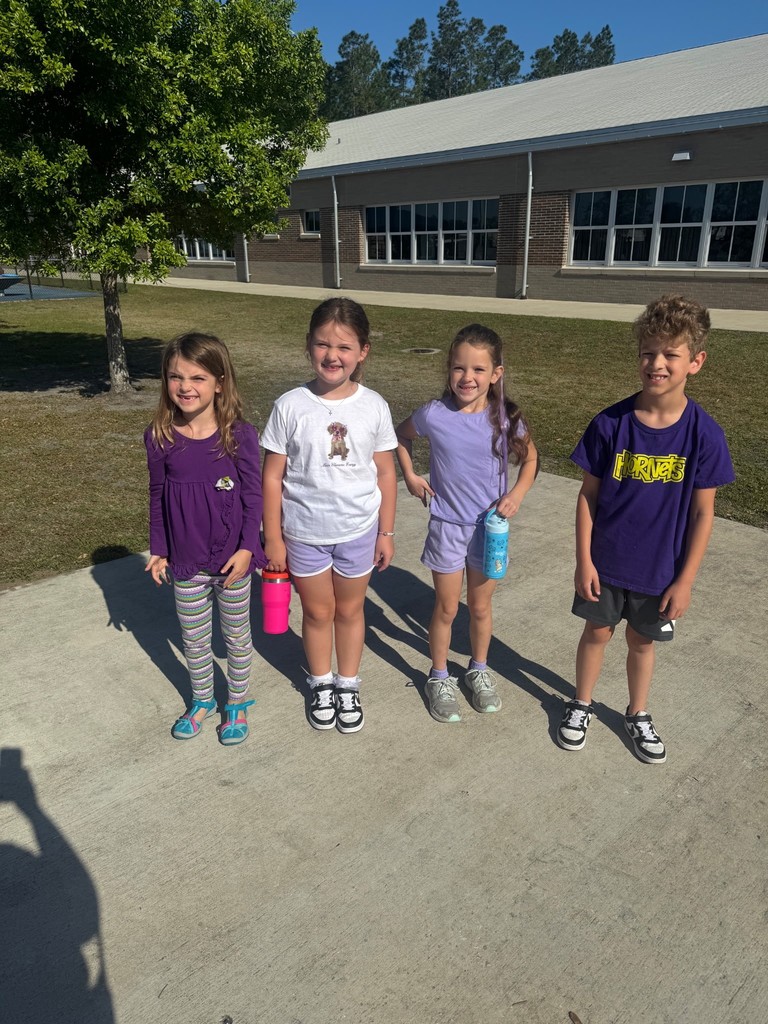 Four elementary students standing together on a sunny sidewalk in front of the school building. They are all wearing purple shirts to show their support for Purple Up Day.