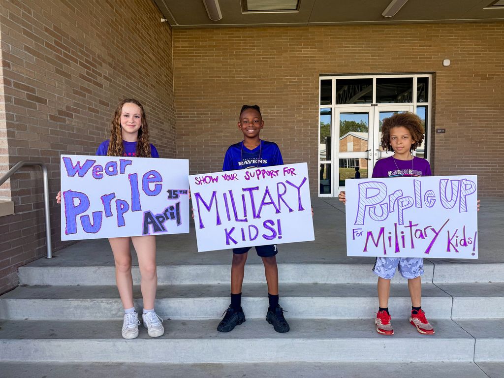 Three elementary-aged students standing on school steps holding handmade signs. The signs say "Wear Purple," "Show your support for Military Kids," and "Purple Up for Military Kids." All three students are wearing purple shirts to celebrate Purple Up Day.