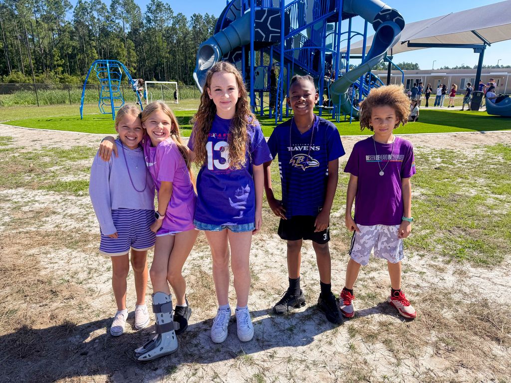 Five elementary students standing together on a sunny school playground, all wearing purple shirts and jerseys to celebrate Purple Up Day. A blue playground structure and green trees are visible in the background.