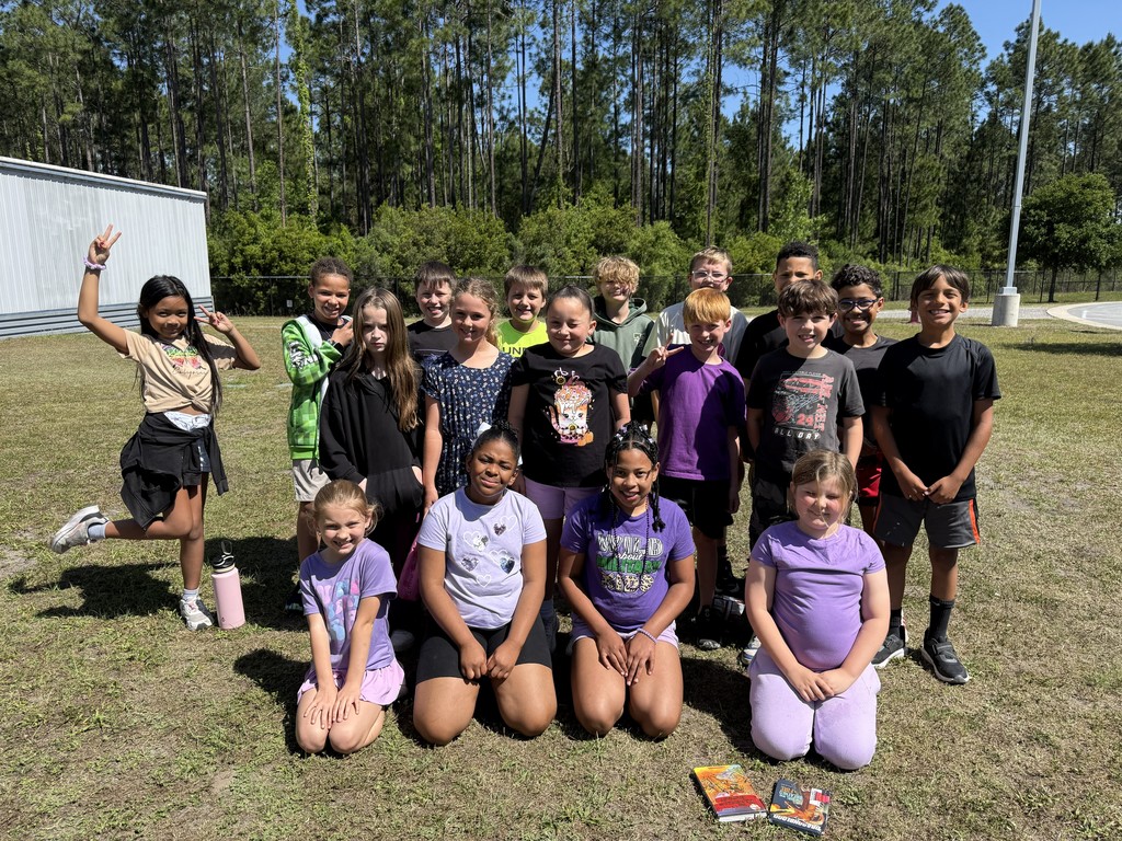 A large group of elementary students posing together on a grassy field outside. Many of the students are wearing purple shirts to celebrate Purple Up Day, with a few sitting in the front and others standing behind them under a clear blue sky.